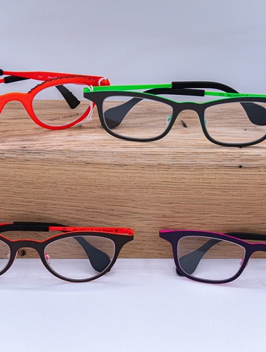 Quatre paires de lunettes modernes aux montures colorées : orange, noir/vert, noir/rouge et violet/rose. Texte visible : 'open rimlock', 'Rim Cup'.